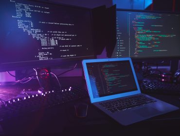 Background image of various computer equipment with programming code on screens on table in dark room, cyber security concept, copy space