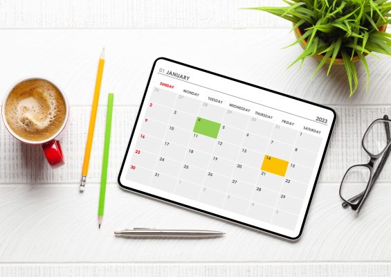 Tablet with calendar app, coffee cup and office supplies on desk. Top view flat lay